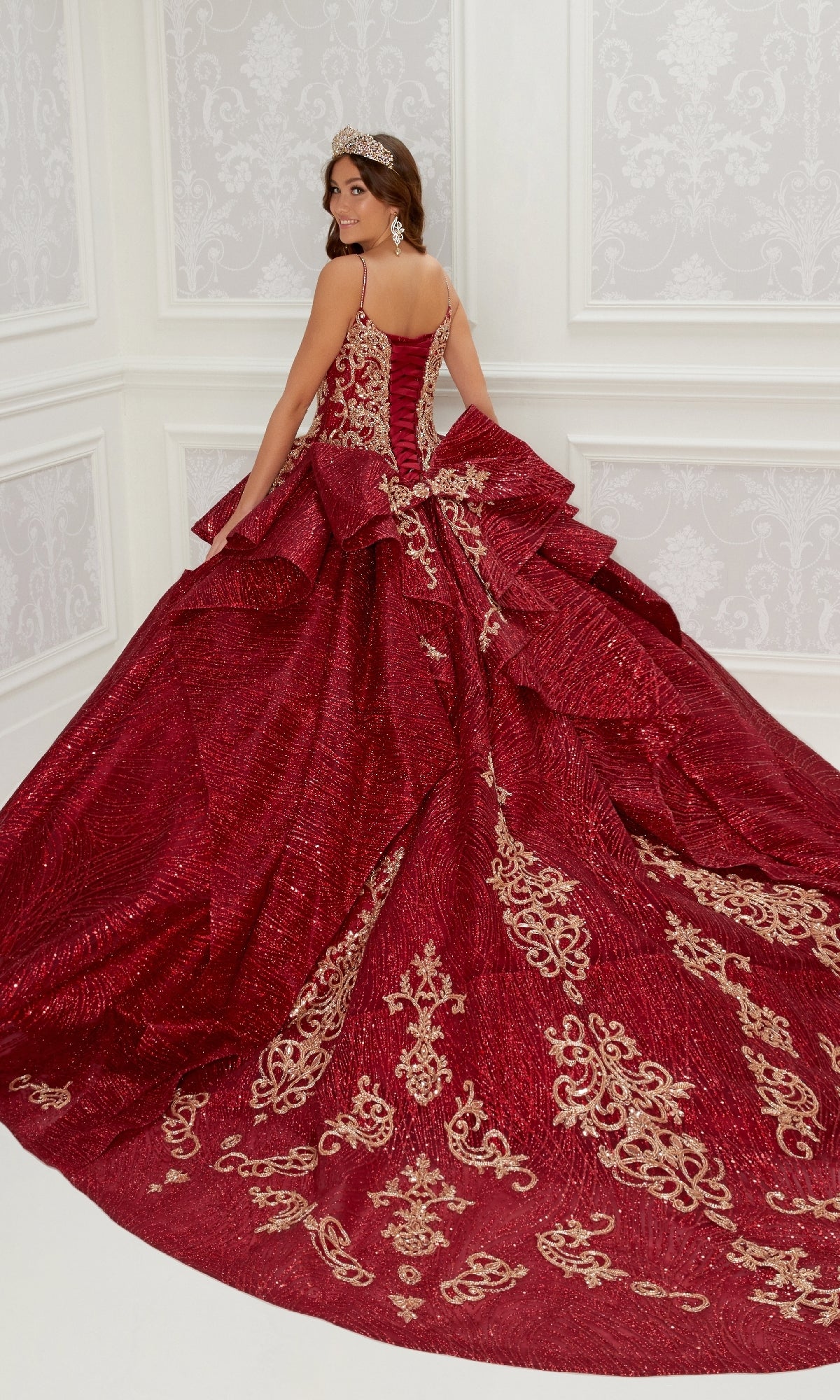A woman wearing the Quinceanera Dress PR22142 By Princesa, featuring deep red fabric, gold embroidery, and a wide train, stands in a decorated white room with her back turned to show off the elegant corset back.
